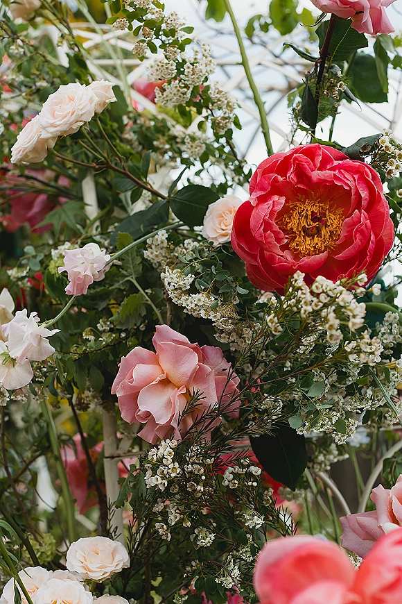 Wedding florals on a ceremony floral arch with pink and coral roses, peonies, baby's breath, and greenery against outdoor sky and foliage.