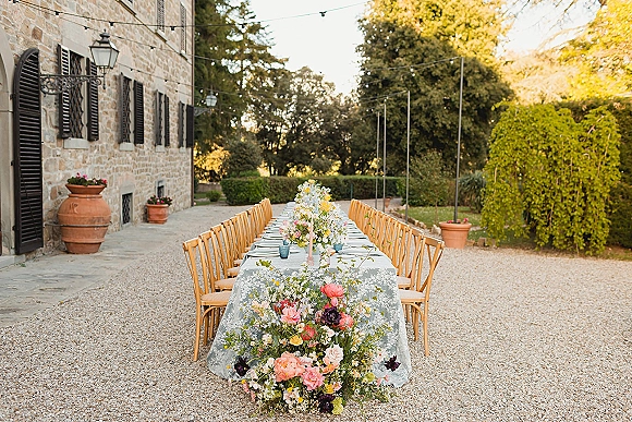 Reception tablescape with long banquet wedding table, lace tablecloth, roses and greenery, taper candles, and string lights in a stone courtyard