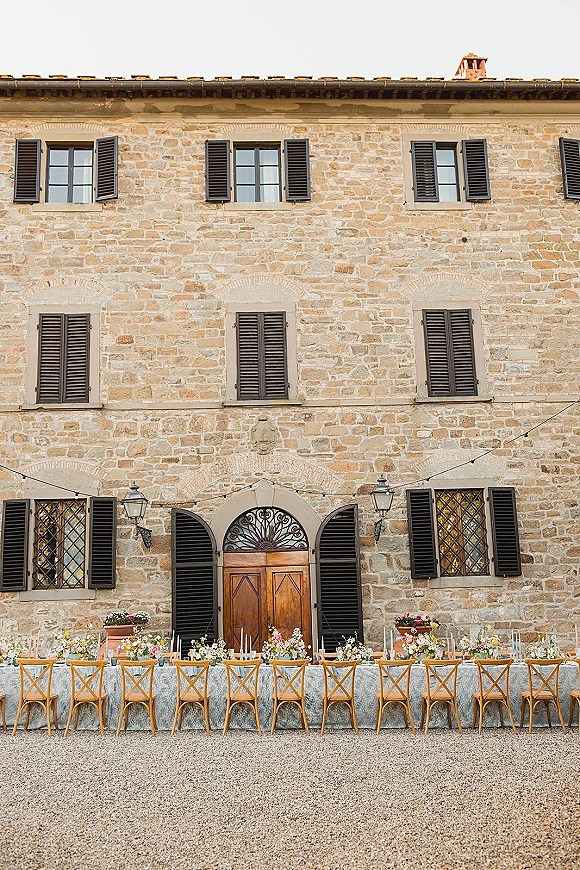 Reception tablescape with a long banquet table, cross back chairs, floral centerpieces and taper candles under string lights in a stone courtyard