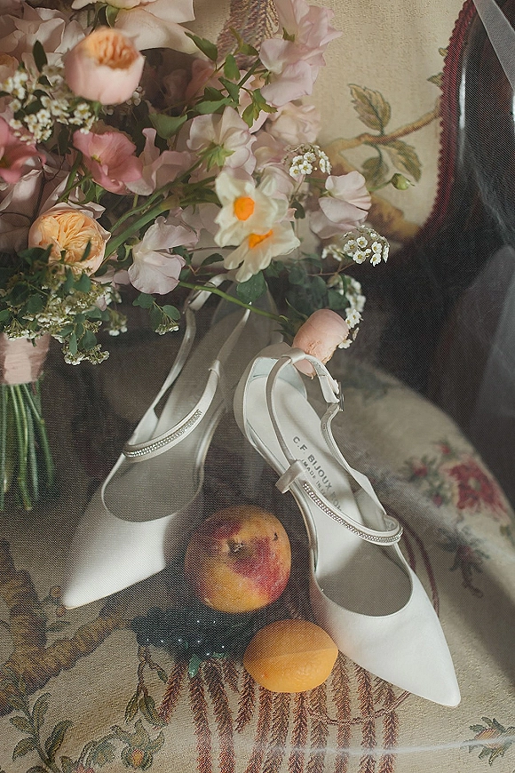 Bridal shoes with white pointed-toe heels and ankle straps, rhinestone trim beside a pastel peach bouquet and veil on floral tapestry fabric