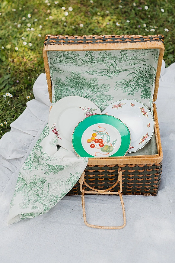 Wedding picnic setup with a wicker picnic basket, patterned napkin, and ceramic plates on a white blanket over a daisy-dotted lawn
