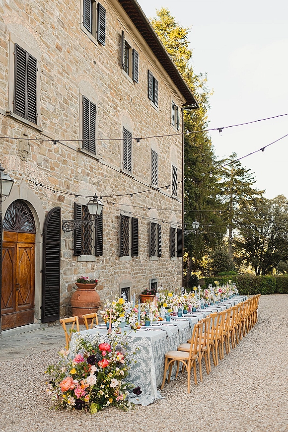 Reception tablescape with long banquet table, blue patterned cloth, wildflower bud vases, taper candles and string lights in a stone villa courtyard