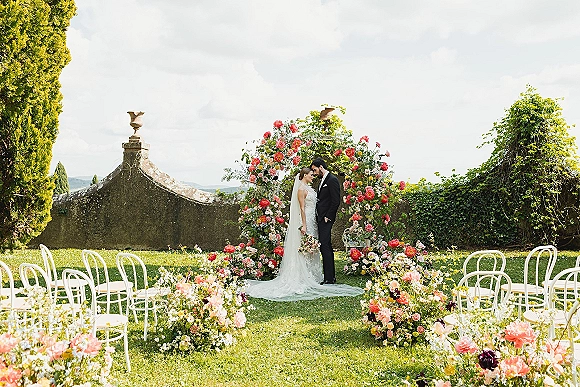 Ceremony moment as bride and groom at altar beneath a floral arch, bouquet and veil on garden lawn by ivy stone wall under clouds