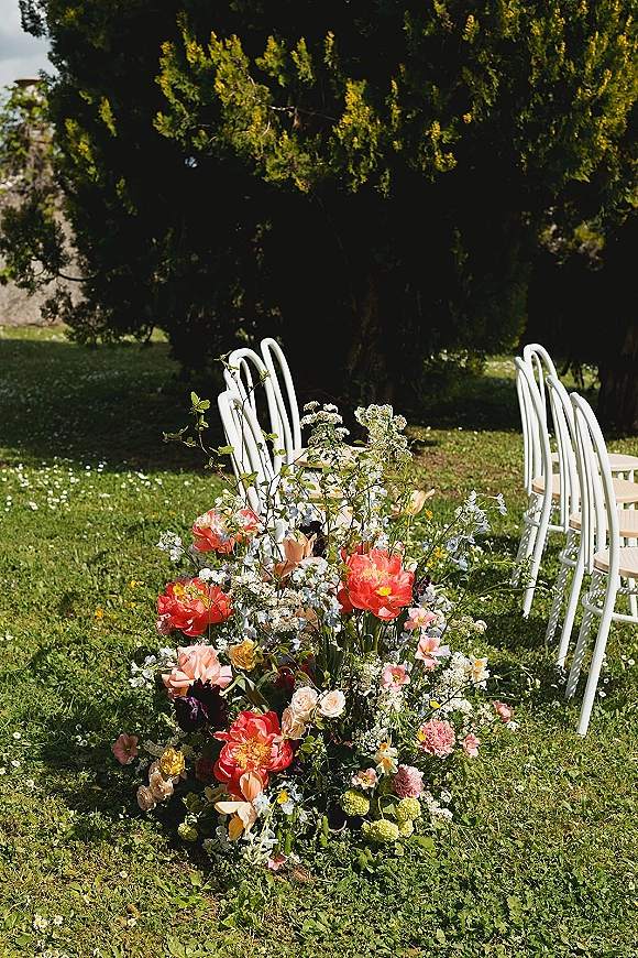 Ceremony aisle florals with coral peonies and wildflowers in a grounded arrangement beside white ceremony chairs on a sunlit lawn under trees
