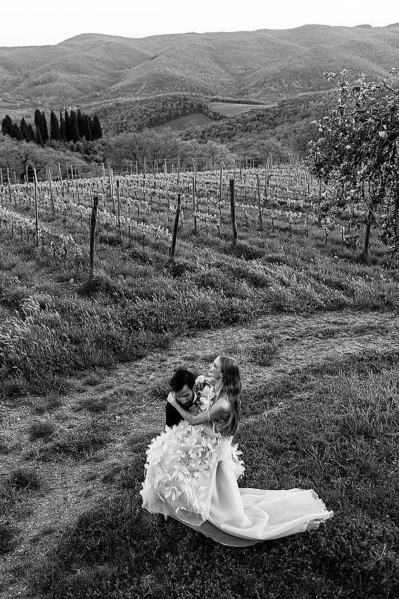 Couple portrait of bride and groom embrace, her long train wedding dress flowing on a dirt path through vineyard hills with mountains behind