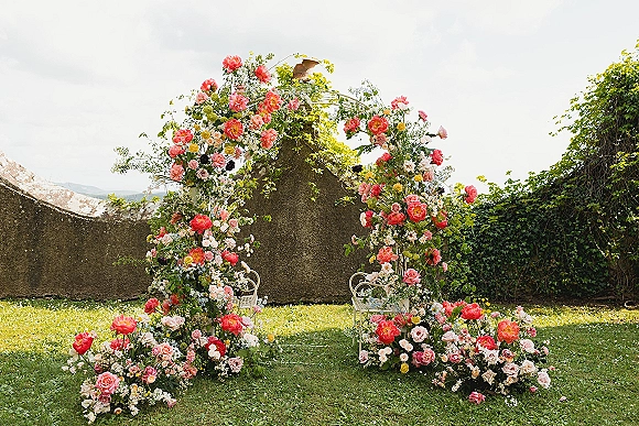 Wedding ceremony arch adorned with pink roses and coral peonies, greenery vines, and meadow base on a lawn beside an ivy stucco wall