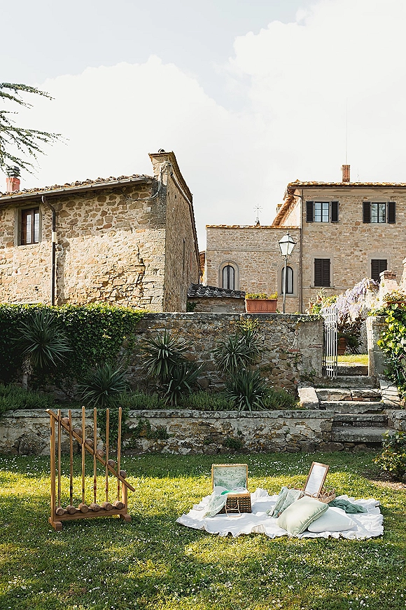 Wedding picnic setup with a wedding picnic blanket, pastel pillows, basket, and wooden lawn game on a garden lawn by stone buildings and gate