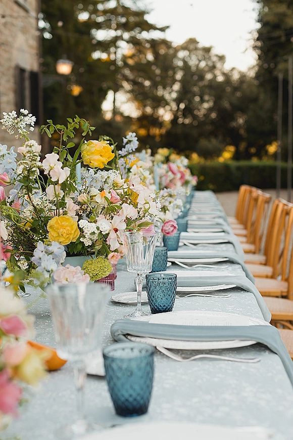 Reception tablescape with pastel floral centerpieces and blue goblets on a long banquet table set on a gravel patio by trees and a stone wall