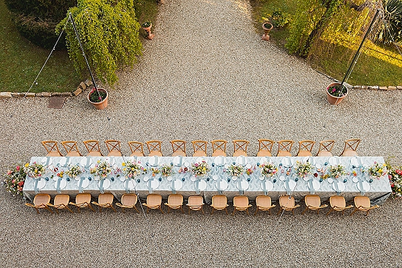 Reception tablescape with a long banquet table setup on blue tablecloth, pastel floral centerpieces and place settings in a gravel courtyard garden