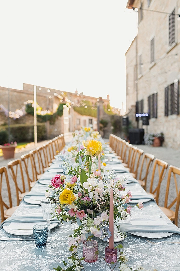 Reception tablescape with long banquet table, floral centerpiece and pink taper candles, colored glass votives under string lights in stone courtyard