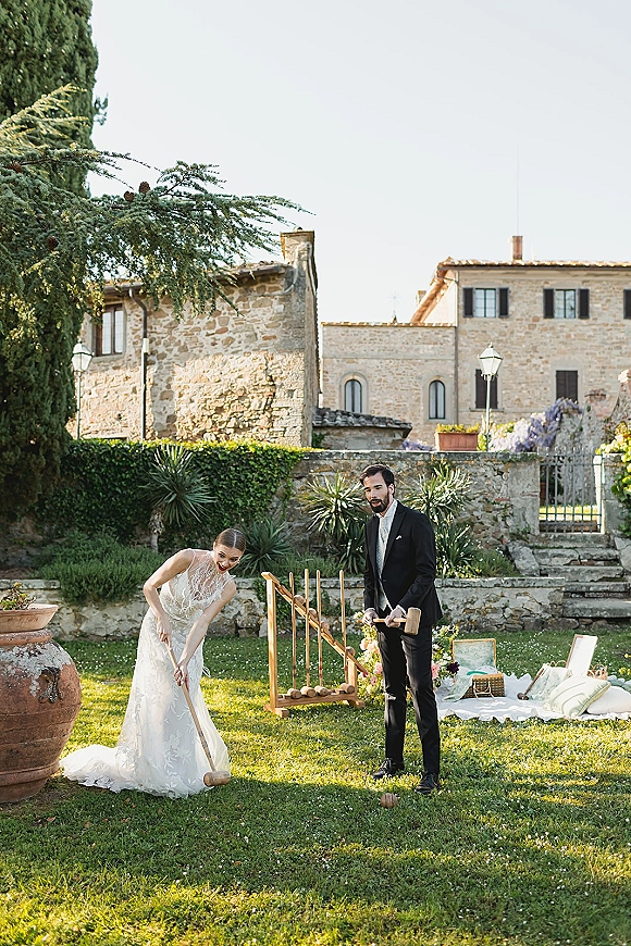 Couple portrait of newlyweds playing croquet, bride in lace wedding dress and groom in black tuxedo on a garden lawn near stone buildings