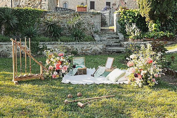 Wedding picnic setup with outdoor wedding lounge area, floral arrangements, blankets and pillows on a grassy lawn by a stone wall and gate
