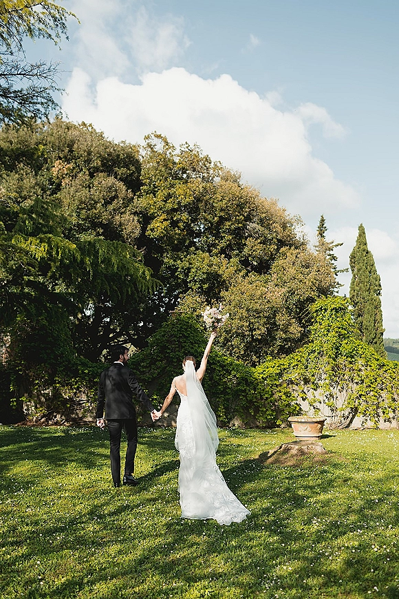 Couple portrait of bride and groom from behind, holding hands as she lifts her bouquet, walking on a garden lawn by a stone wall
