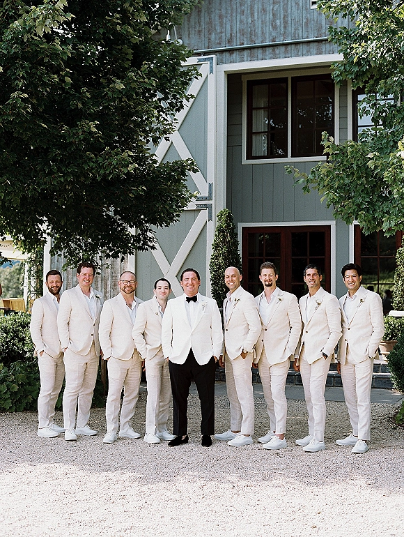 Groomsmen portrait of the groom with groomsmen in cream suits with boutonnières, black bow tie and white sneakers outside a barn with trees