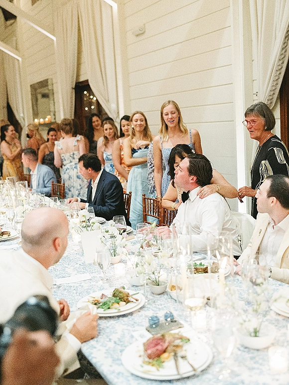 Wedding reception toast as maid of honor gives a speech beside a long blue patterned banquet table with taper candles and listening guests