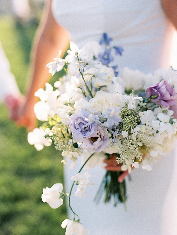 Bridal bouquet of white and lavender bouquet flowers with greenery accents, held against a soft outdoor lawn backdrop