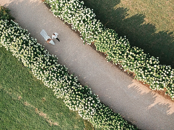 Wedding couple portrait from above, bride in long gown and veil holding groom’s hand on a gravel garden path lined with hydrangeas