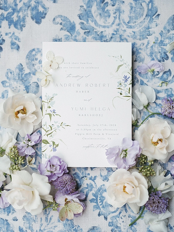 Wedding invitation with watercolor floral invitation details, white and lavender flowers and greenery on blue patterned linen backdrop flat lay