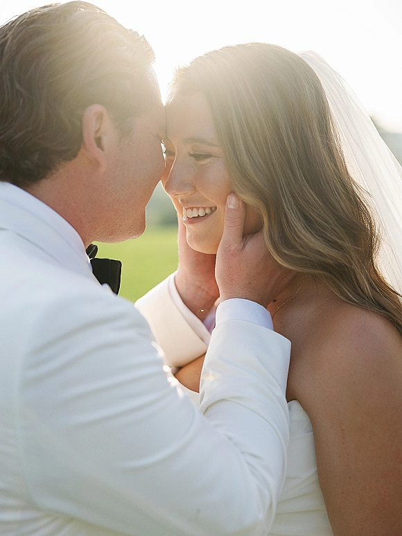 Wedding couple portrait with bride and groom close up, forehead touching as he cups her face, sunset backlit in a grassy field with veil
