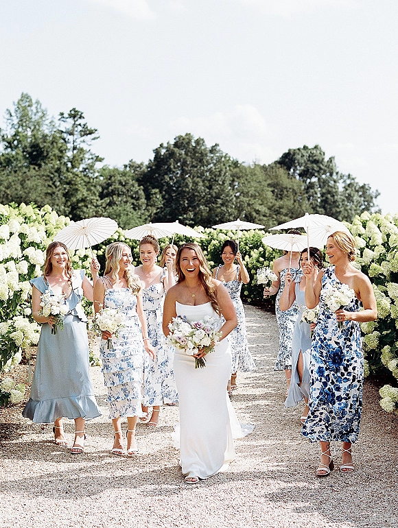 Bride with bridesmaids walking photo along a garden path, laughing as they carry bouquets and a white paper parasol by hydrangeas