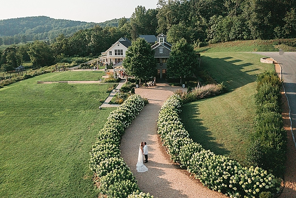 Couple portrait from above of bride in long veil and wedding dress beside groom in suit on a hydrangea-lined gravel driveway at a farmhouse estate