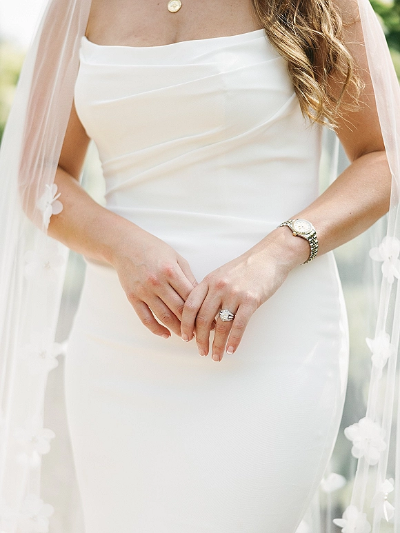Bridal dress details with a strapless wedding dress, veil draped over the shoulders as hands show an engagement ring against greenery
