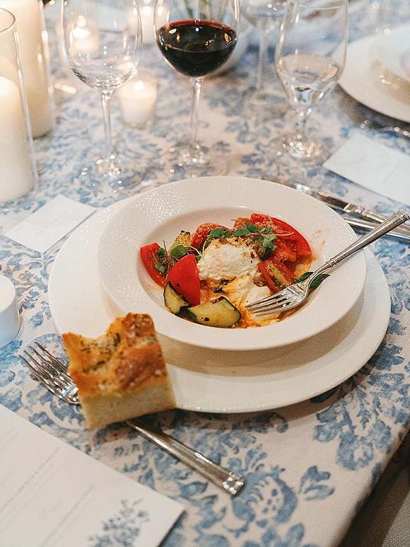 Wedding dinner table with wedding place setting, white porcelain dinnerware, red wine glasses, and candles on a blue toile tablecloth
