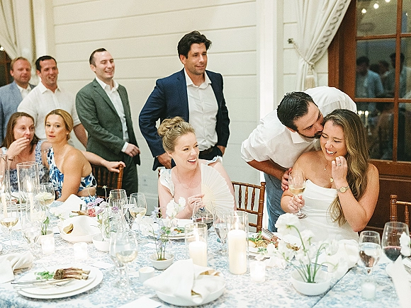 Reception toast moment as the bride laughs while the groom kisses her cheek at a candlelit sweetheart table in an indoor room with guests