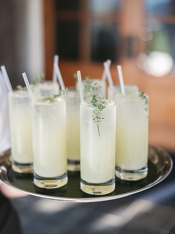 Signature cocktails in highball glasses with clear straws and herb garnish on a serving tray, set against a blurred indoor reception space