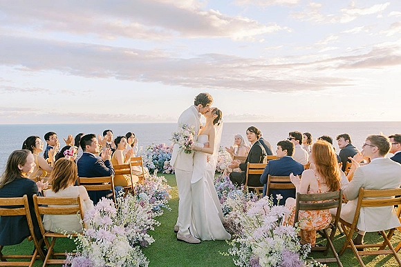 Ceremony kiss as bride in long veil holds bouquet, kissing groom in white suit on a lawn aisle with flowers by the ocean horizon