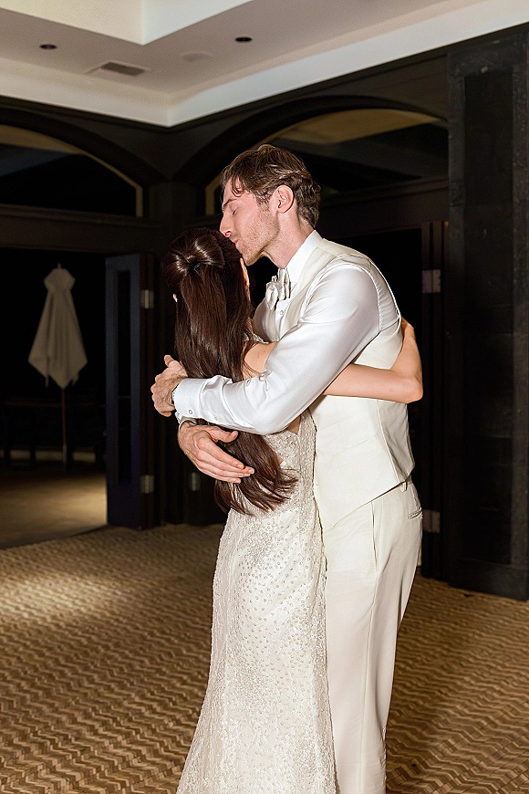 First look moment as groom kisses bride’s forehead in a wedding first look hug, her beaded dress sparkling in a dark-wood arched hallway