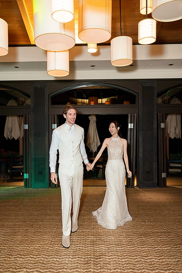 Newlywed entrance as bride and groom walking hand in hand, her beaded halter gown and his vest under pendant lights by an arched doorway