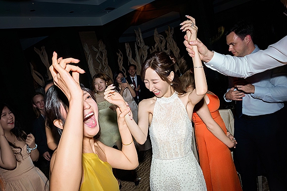 Wedding dance floor packed with bride in a beaded gown holding hands and laughing with guests during an indoor reception dance party