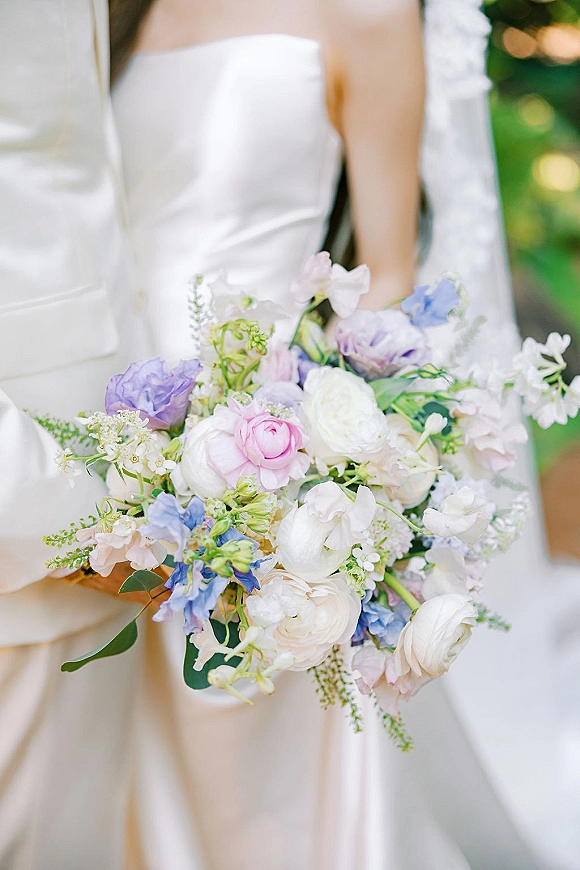 Bridal bouquet of pastel wedding bouquet blooms with pink rose, white ranunculus and blue delphinium, held against a strapless dress with greenery bokeh