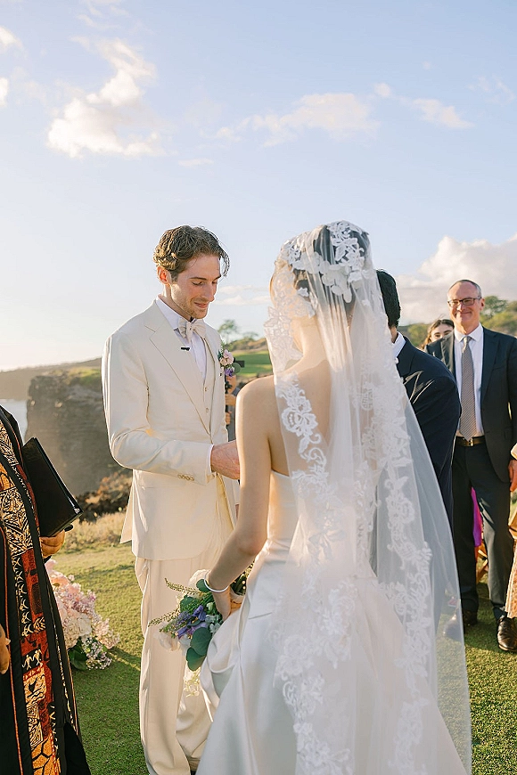 Wedding vows as bride in lace veil and gown holds purple-white bouquet, groom in cream suit by microphone on hillside lawn under blue sky