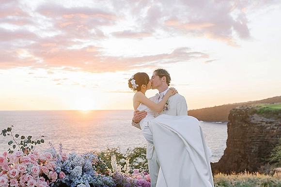 Wedding kiss portrait of groom carrying bride in a white dress and suit, hair flowers visible, with ocean cliffs and sunset clouds behind