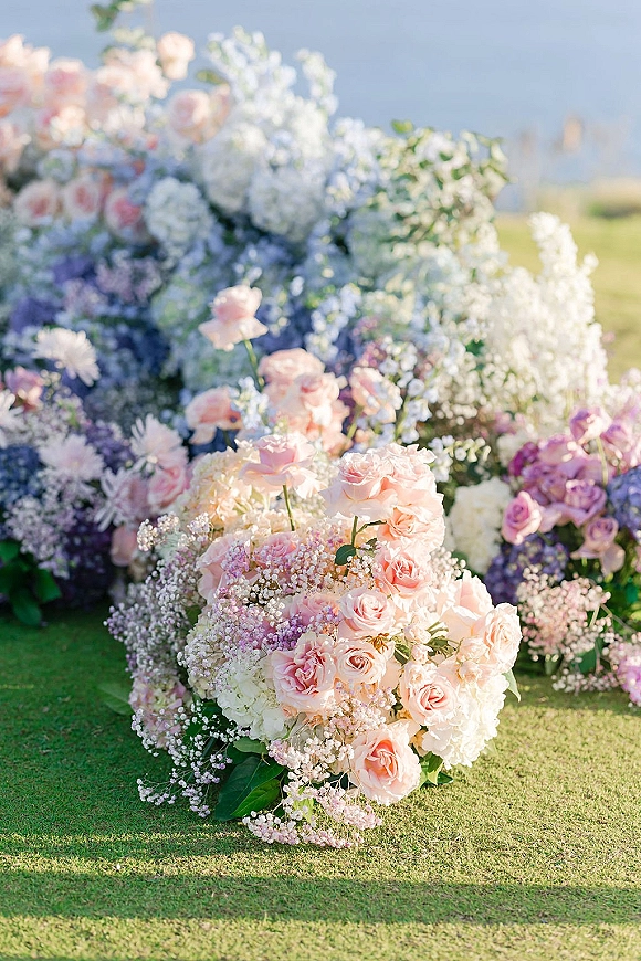 Wedding floral arrangements with blush roses accent in low aisle clusters on a grass lawn beside ocean water under open sky