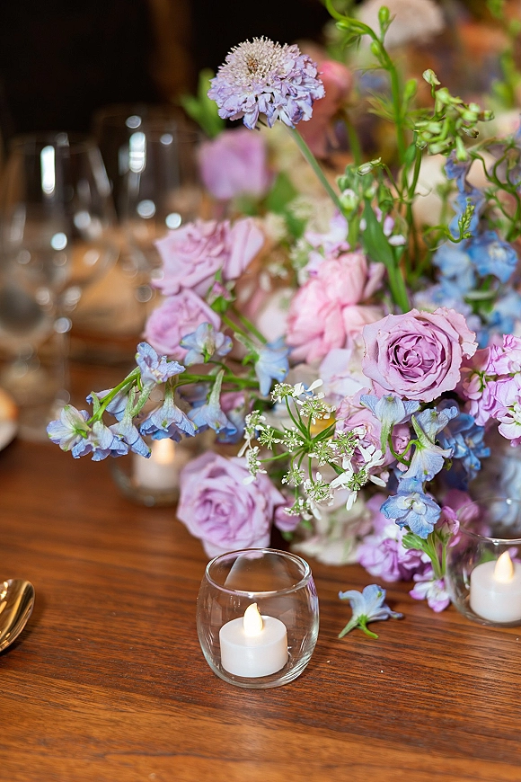 Reception centerpiece of pastel wedding centerpiece blooms with lavender roses, blue delphinium, and candles on a wooden table in warm light