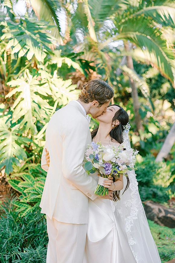 Wedding kiss portrait of bride and groom kissing, bride holding a pastel bouquet with long lace veil amid tropical garden greenery