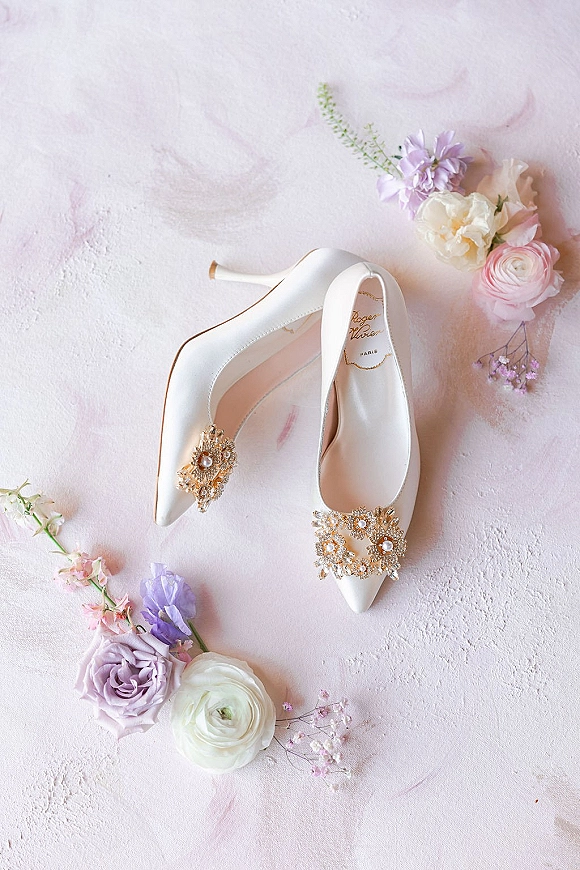 Wedding shoes in white satin heels with jeweled shoe clips, pearl and gold accents, styled with pastel flowers on a pink backdrop