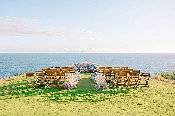 Ceremony setup with wood folding chairs and floral aisle arrangements facing an asymmetrical altar on a grassy cliff with ocean view