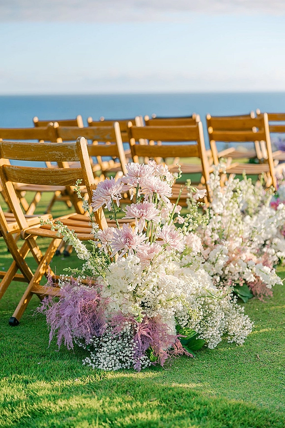 Ceremony aisle flowers in pink and white with baby’s breath and greenery beside wood folding chairs on a seaside lawn with ocean view