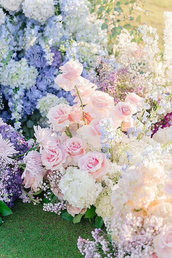Wedding floral arrangement of pastel wedding flowers with pink roses, hydrangeas, lilac blooms, and greenery on a sunlit grass lawn