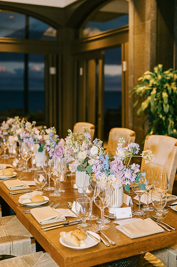 Reception tablescape with wood banquet table setting, pastel flower centerpieces in ribbed white vases, menus, place cards, and glassware by glass doors