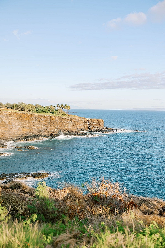 Ocean cliff view at a coastal wedding venue overlook, with waves on rocky shoreline, palm trees, blue sky, and a distant island silhouette
