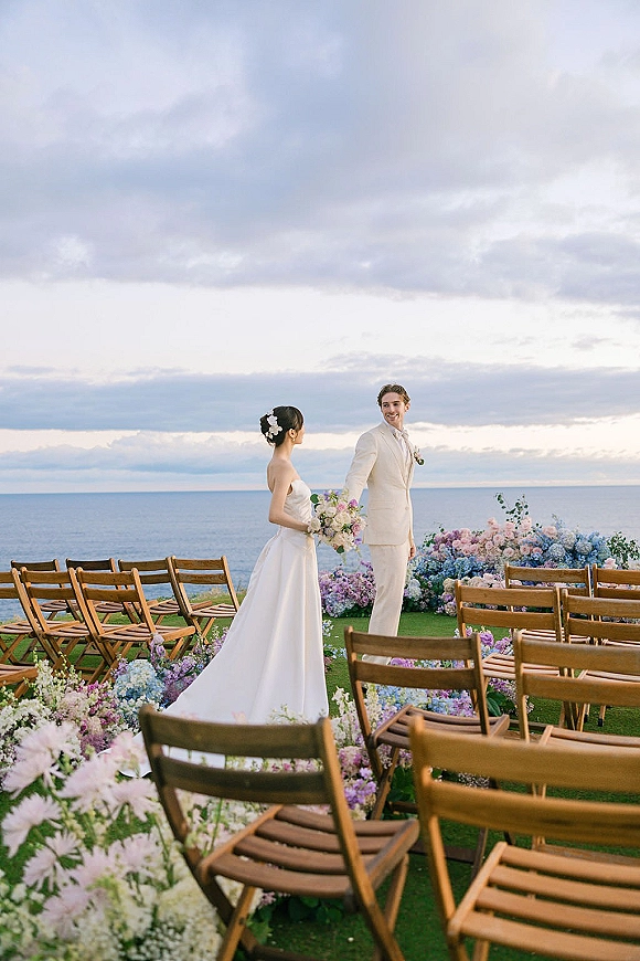 Ceremony moment at an outdoor oceanfront ceremony with bride and groom holding hands by aisle florals, wooden chairs, and ocean horizon backdrop