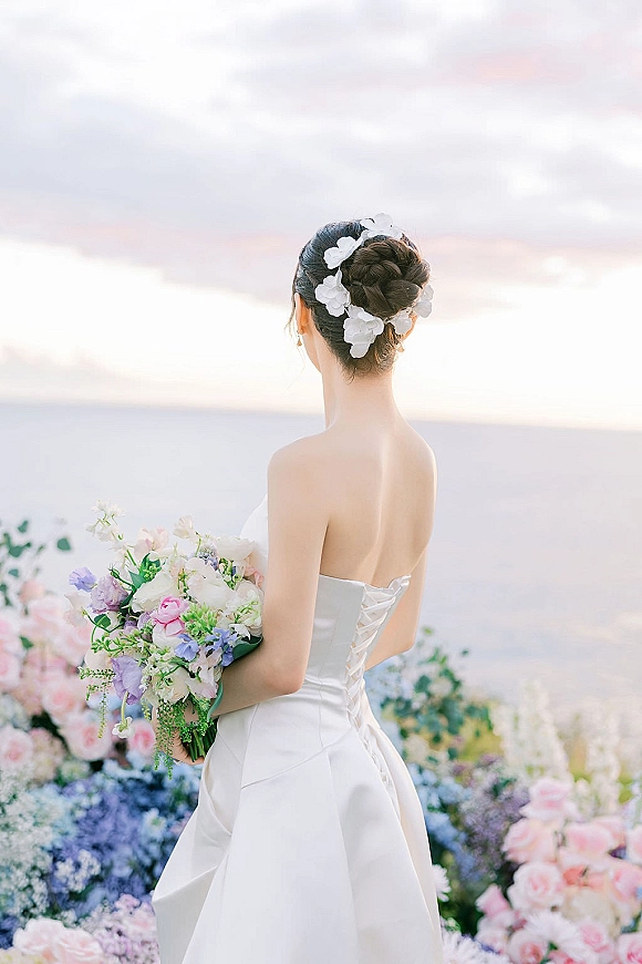 Bridal portrait of a bride from behind holding a pastel bouquet, strapless corset-back gown and floral updo against an ocean sunset horizon