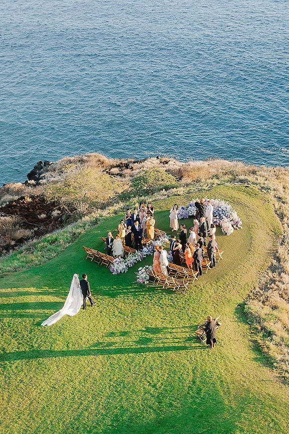 Outdoor wedding ceremony with bride in flowing veil walking to groom beneath a floral arch on a cliffside lawn above the ocean