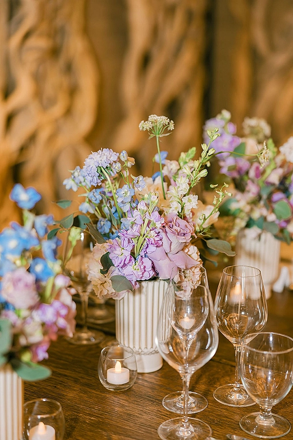 Reception tablescape with a wedding table centerpiece of pastel flowers in white ribbed ceramic vases, tea lights, and stemware against gold drapery