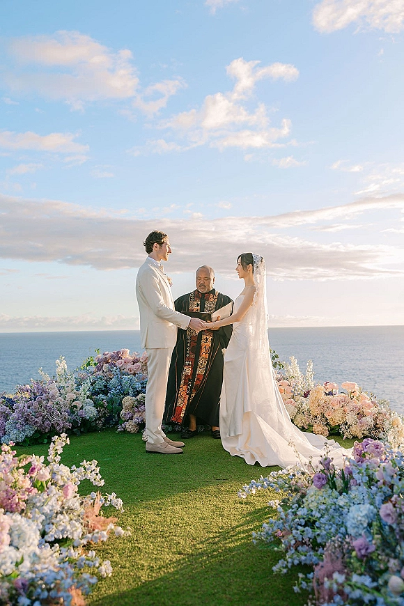 Wedding vows during an outdoor wedding ceremony as the bride and groom hold hands, bride’s veil flowing before an ocean backdrop